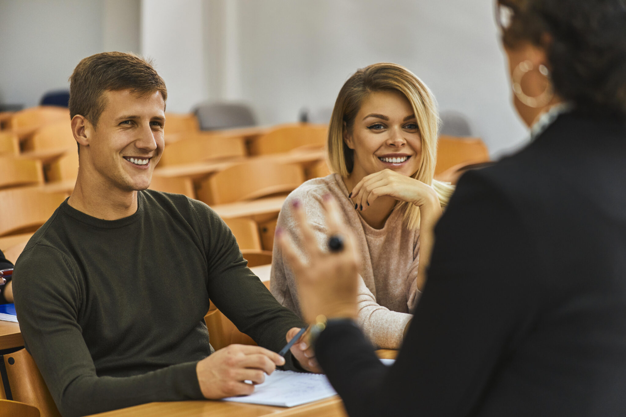Two smiling students sitting in an auditorium, attentively listening to a lecturer